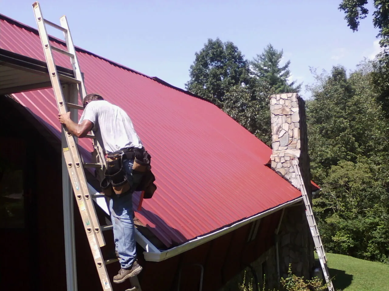 Roofer on a ladder beside a red metal roof and stone chimney — Soffit & Fascia Repair by Roofing Brothers in Fair Bluff